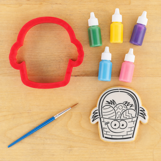 Easter Basket Paint Your Own Cookie lying next to the matching Easter Basket Cookie Cutter and bottles of pastel food coloring and a food grade artist brush.