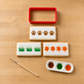A flat-lay of a sugar cookie decorating kit on a light wood background, featuring a red rectangular cookie cutter, three decorated cookies with paw prints, grass, and flowers, a paintbrush, and a paint palette.