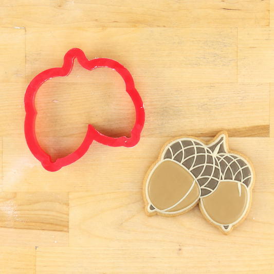 Acorn shaped cookie cutter, next to an acorn shaped cookie that has been decorated using the Acorn Cookie Stencil.