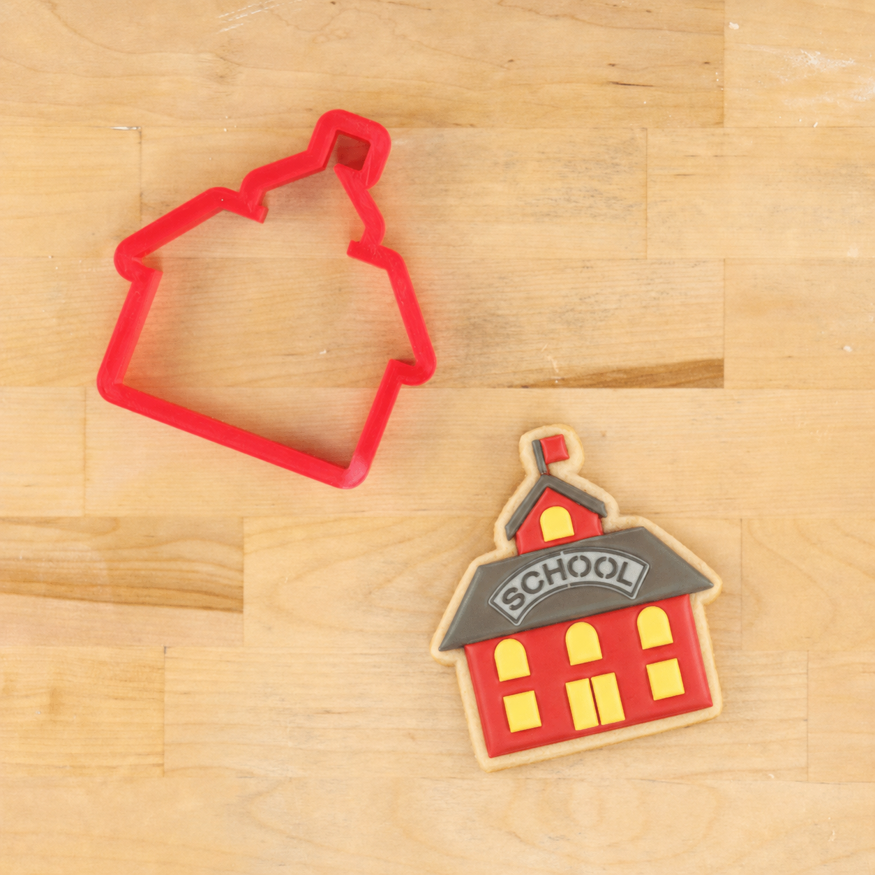 A red 3D Printed, plastic cookie cutter shaped like a schoolhouse,  shown on a light wood surface next to a decorated sugar cookie in the same schoolhouse shape with red icing, yellow windows, and a gray roof with the word 'SCHOOL' on it.