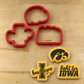 Four red cookie cutters arranged on a light wood surface, with three matching yellow-and-black Iowa Hawkeyes-themed cookie cutters or cookie shapes placed below them.