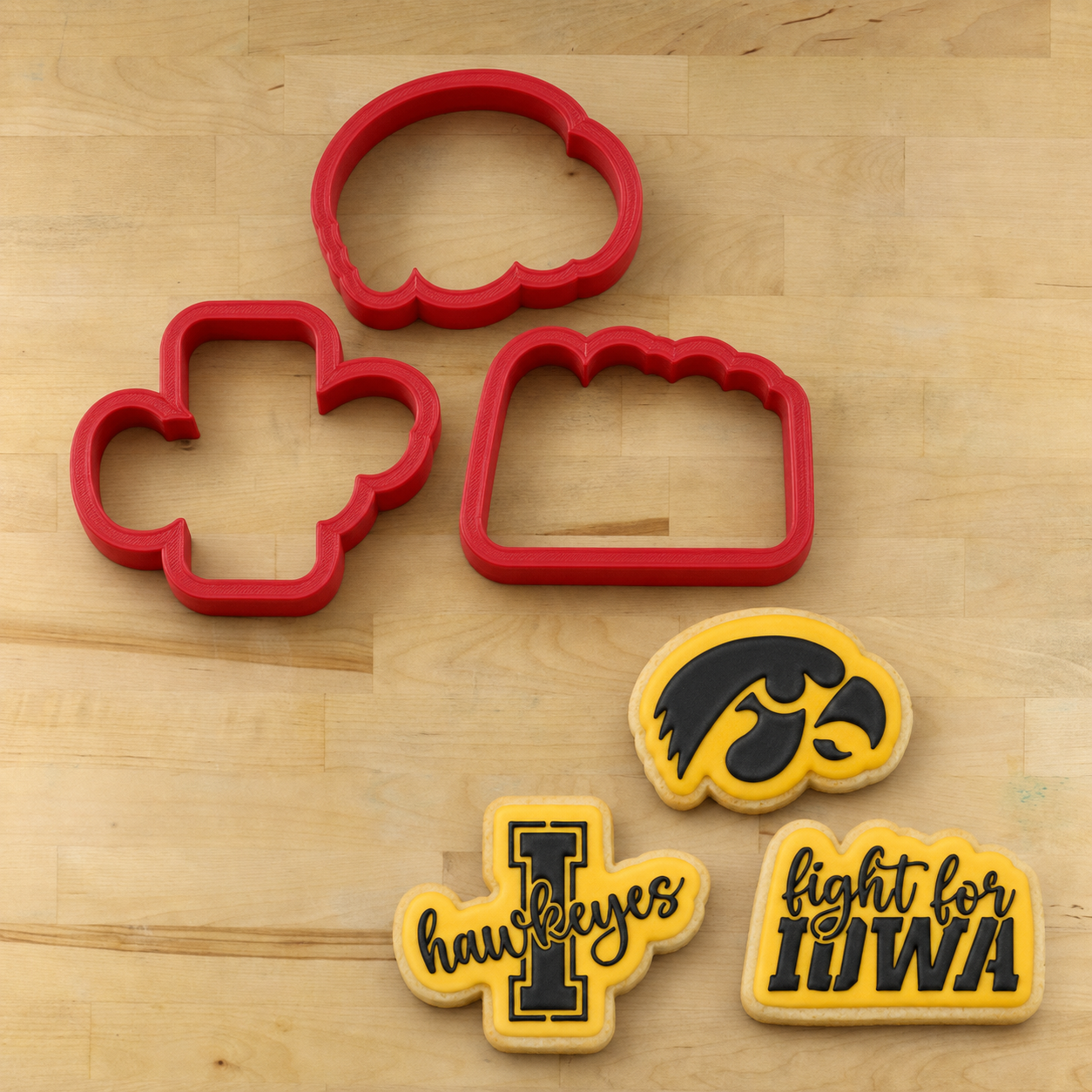 Four red cookie cutters arranged on a light wood surface, with three matching yellow-and-black Iowa Hawkeyes-themed cookie cutters or cookie shapes placed below them.