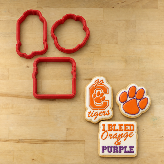 A set of red cookie cutters and three decorated sugar cookies on a light wood background. The cookies feature Clemson Tigers branding, including the tiger paw logo and 'I Bleed Orange & Purple' text.