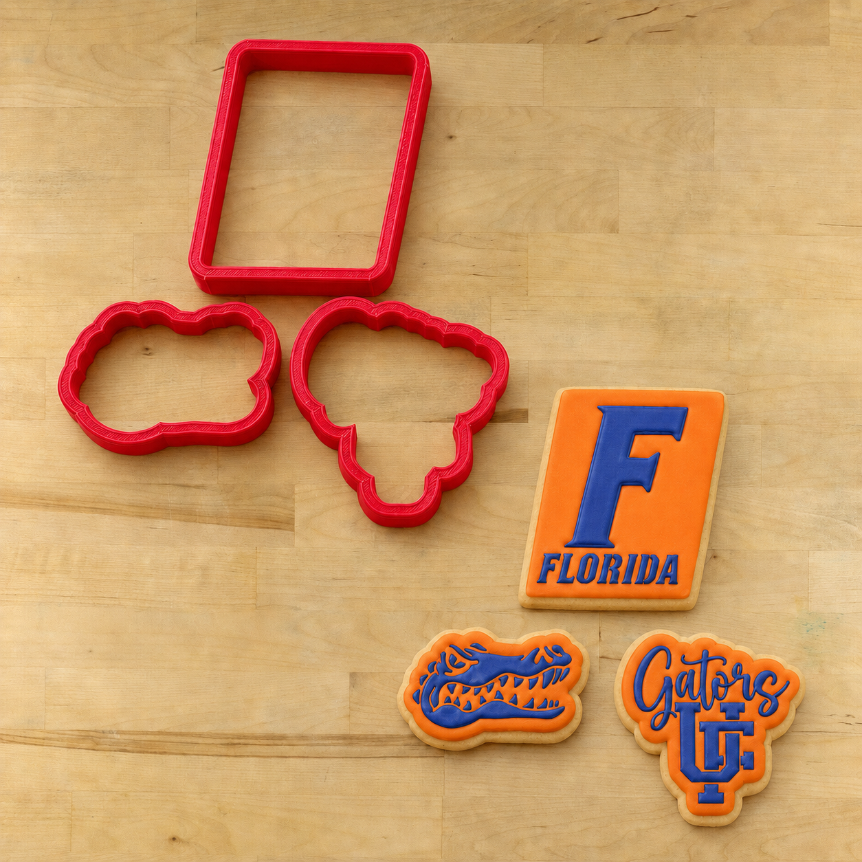 Red plastic cookie cutters and decorated sugar cookies on a light wood surface. The set includes a rectangular cutter, two cloud-shaped cutters, and three decorated cookies featuring the Florida Gators logo in orange and blue icing.