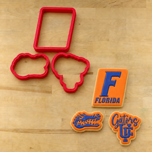 Red plastic cookie cutters and decorated sugar cookies on a light wood surface. The set includes a rectangular cutter, two cloud-shaped cutters, and three decorated cookies featuring the Florida Gators logo in orange and blue icing.