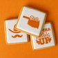 Three square sugar cookies with white icing and orange Oklahoma State decoration, arranged on a bright orange fabric background. 