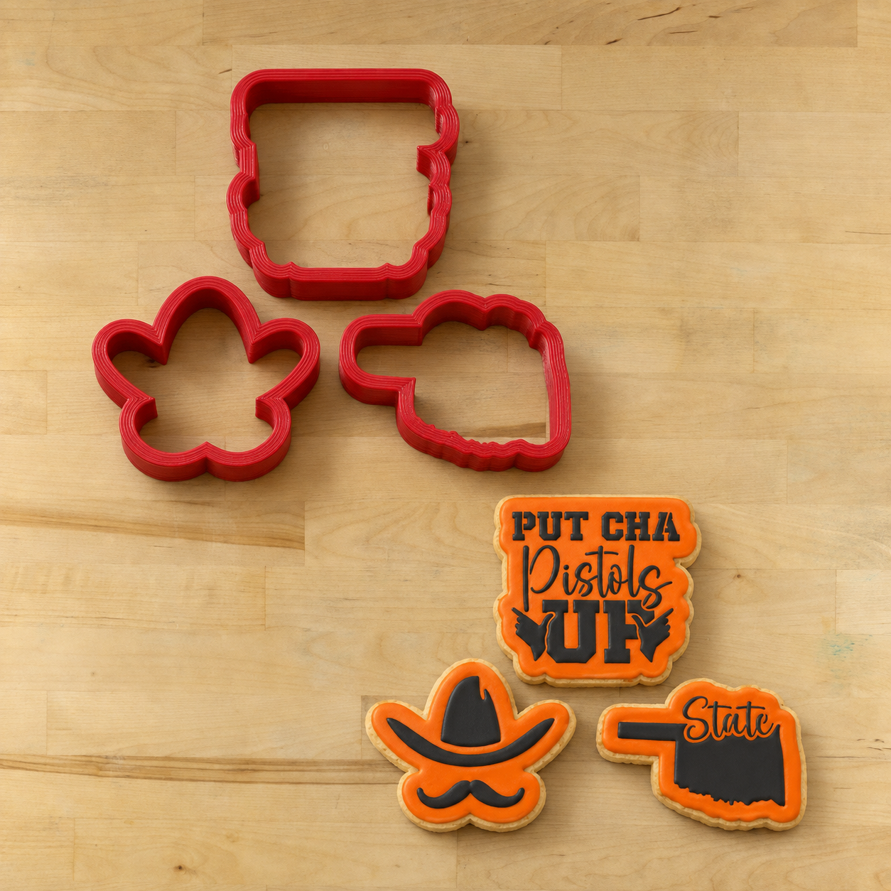 Three red cookie cutters arranged on a light wood surface next to three decorated cookies with Oklahoma State Football Mottos on them 