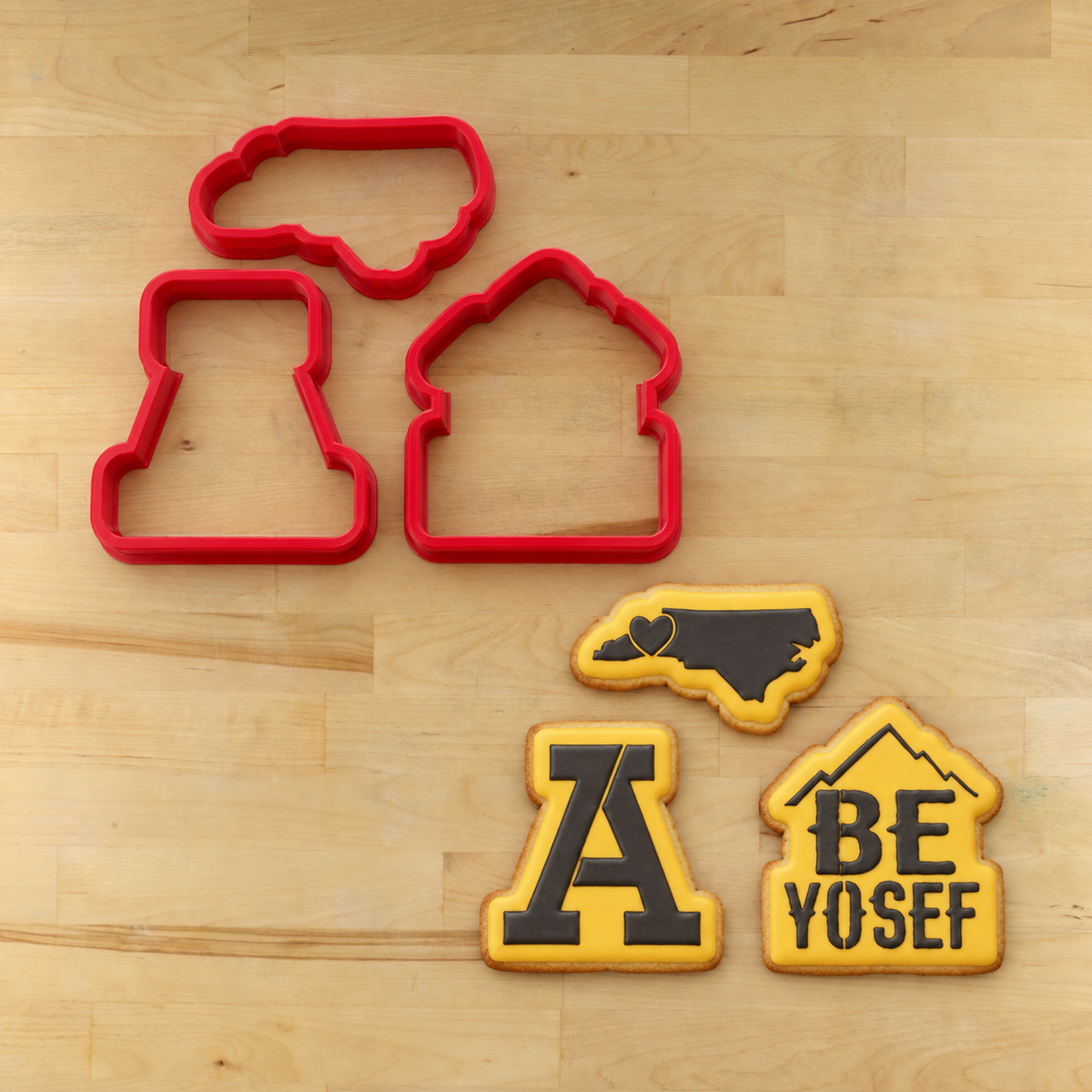 Three red cookie cutters on a light wood surface, with three decorated cookies shown below: a North Carolina cookie, an 'A' cookie, and a house-shaped cookie that reads 'BE YOSEF'.