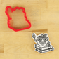 One Smart Apple Cookie stencil, applied to a white, iced cookie lying next to the matching cookie cutter.