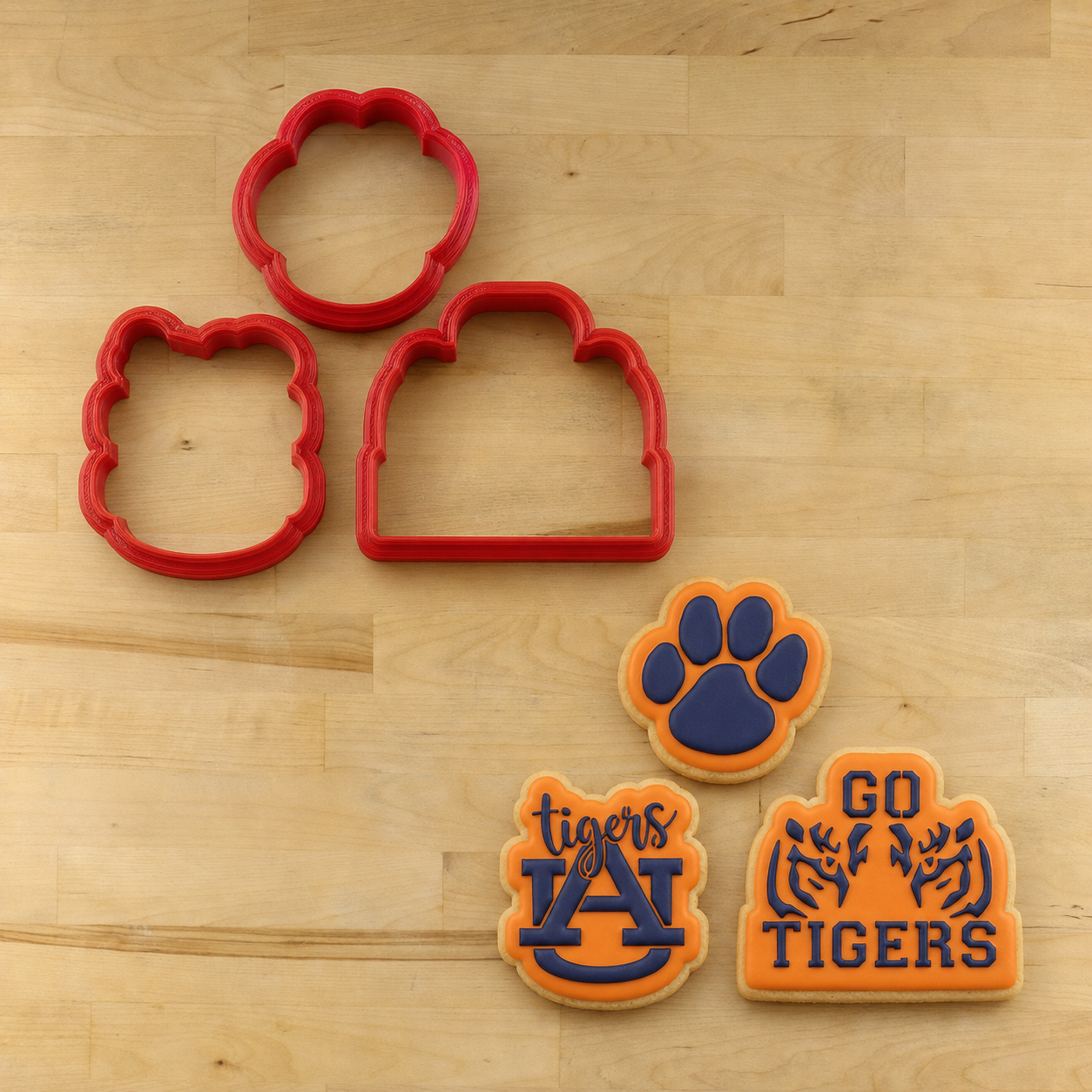 Three red cookie cutters and three decorated cookies shaped like a paw print, house, and Auburn Tigers logo, arranged on a light wood surface.