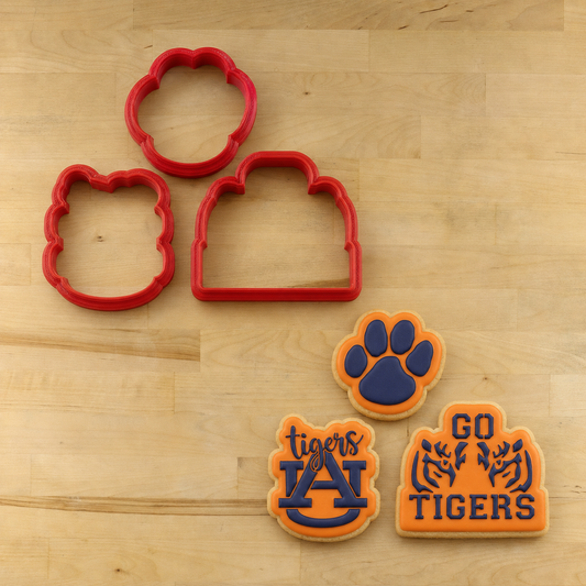 Three red cookie cutters and three decorated cookies shaped like a paw print, house, and Auburn Tigers logo, arranged on a light wood surface.