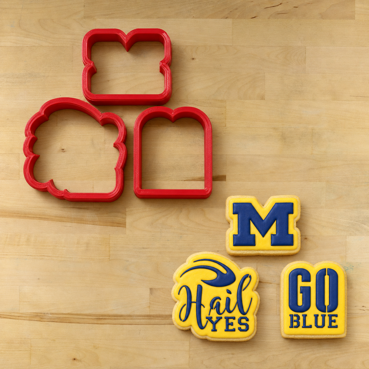 Michigan Wolverines Cookie Cutters lying next to three cookies decorated with the matching "Hail Yes" Cookie Stencil Set