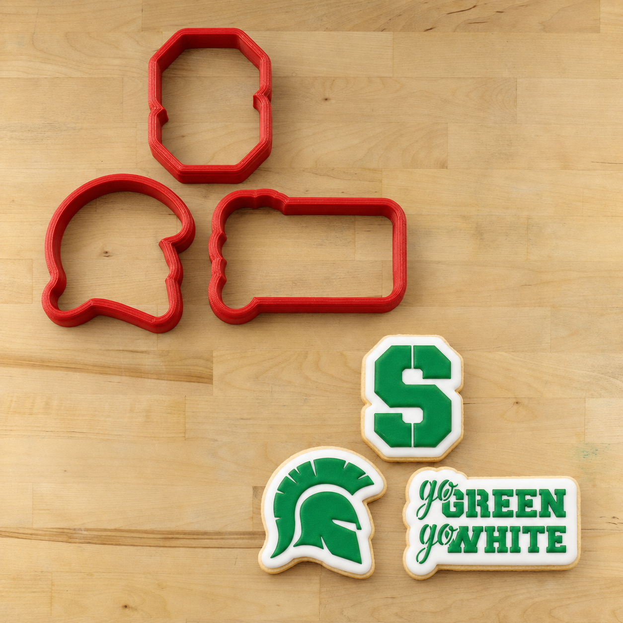 Four red cookie cutters arranged on a light wood surface, with three decorated sugar cookies shown below them featuring the Michigan State Spartans logo and the phrase 'go GREEN yo'WHITE'.