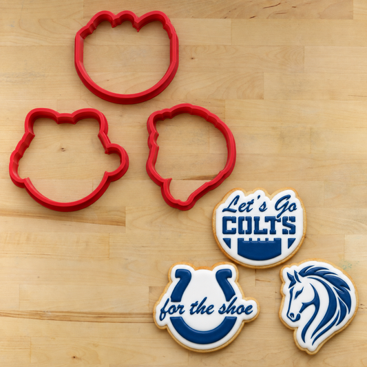 Three red cookie cutters shaped like the Indianapolis Colts horse head logo are arranged on a light wood surface next to three decorated cookies featuring the Colts logo in blue and white.