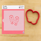 A pink cookie stencil sheet with two red holiday-themed footprints reading 'HOHO HO' and 'MERRY MOO' with snowflakes and trees, shown next to a red heart-shaped cookie cutter on a light wood background. The top of the stencil card reads 'Cookie Stencils' and shows the 'Confict Craze' brand logo.