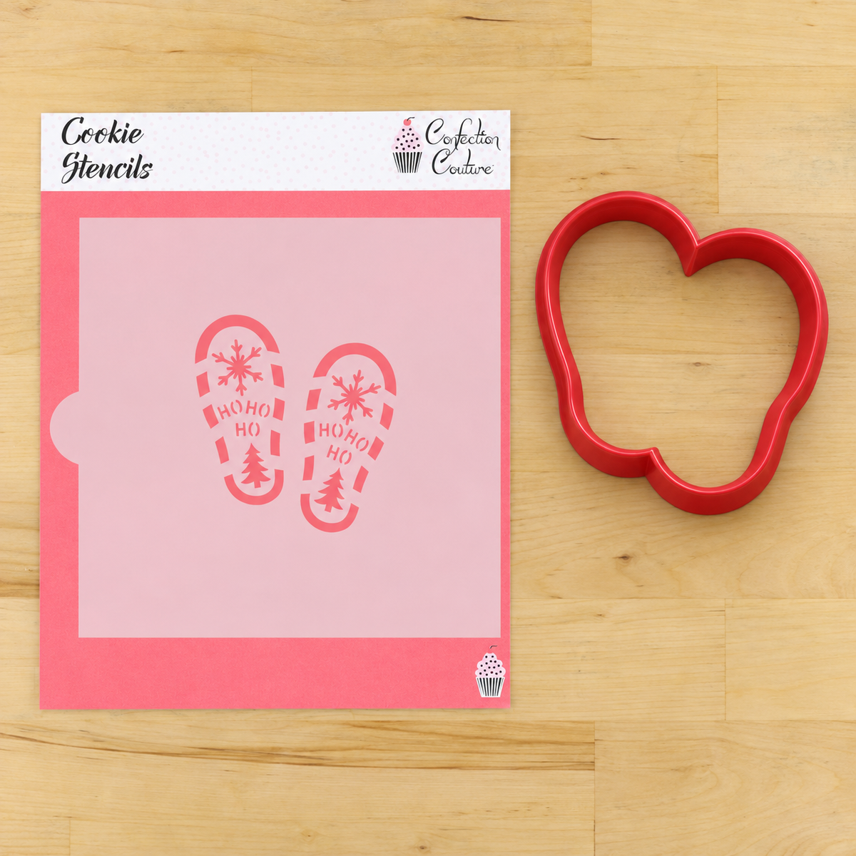A pink cookie stencil sheet with two red holiday-themed footprints reading 'HOHO HO' and 'MERRY MOO' with snowflakes and trees, shown next to a red heart-shaped cookie cutter on a light wood background. The top of the stencil card reads 'Cookie Stencils' and shows the 'Confict Craze' brand logo.