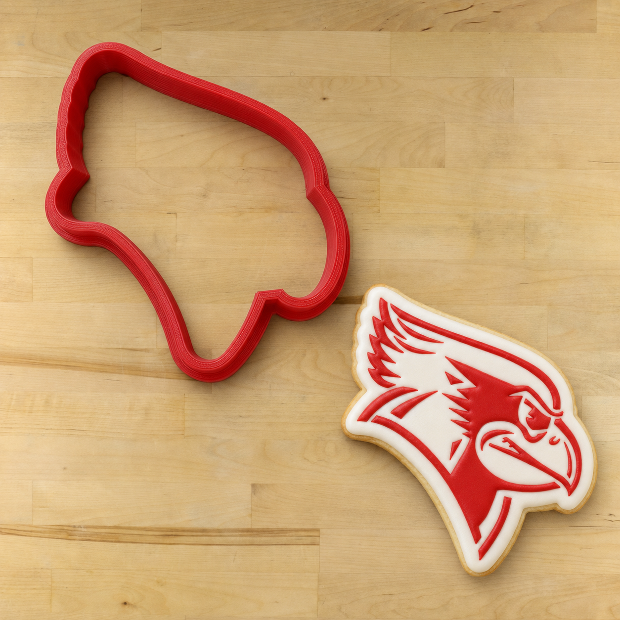 A red cookie cutter shaped like a cardinal head is shown on a light wood surface next to a decorated sugar cookie in the same cardinal head design.