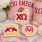 Three decorated Chi Omega sugar cookies displayed with pink sorority-themed décor, ribbons, and floral accents.