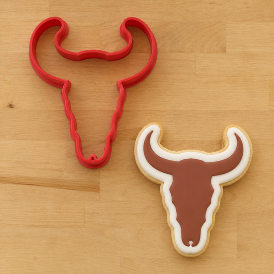 A red bull skull-shaped cookie cutter placed on a light wood surface next to a decorated sugar cookie in the same shape, showing the outline of the cutter and a sample of the cutout cookie.