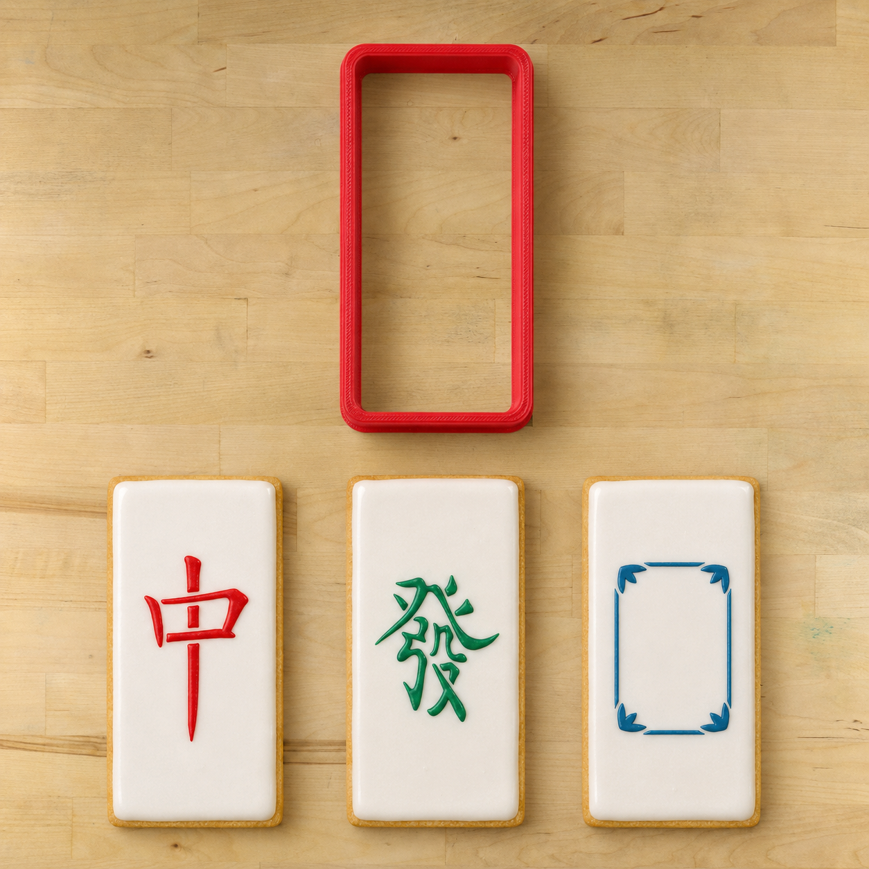 Three white rectangular cookies with Chinese mahjong-style symbols and a red rectangular cookie cutter, arranged on a light wood surface.