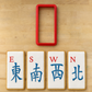 A red rectangular cookie cutter is shown above four decorated sugar cookies with white icing, gold edges, and blue Chinese characters for compass directions, arranged on a light wood surface.