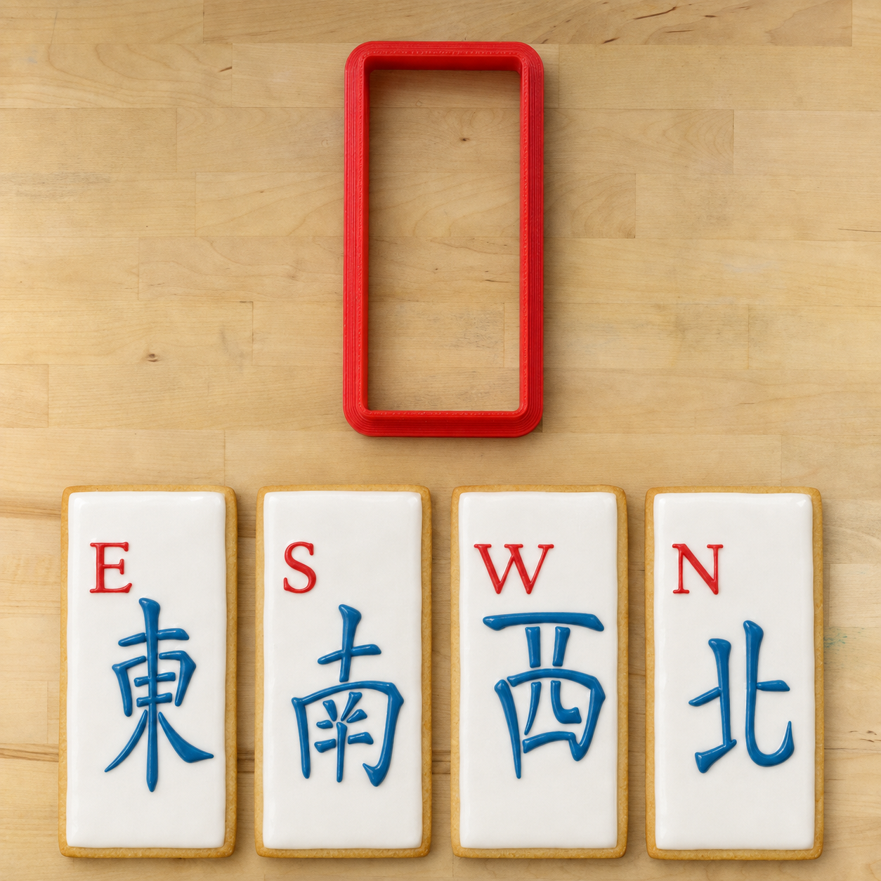 A red rectangular cookie cutter is shown above four decorated sugar cookies with white icing, gold edges, and blue Chinese characters for compass directions, arranged on a light wood surface.