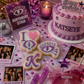 A styled birthday party scene with pink and purple decorations, a custom cake reading KATSEYE, cookie cutouts shaped like letters and an eye, printed photo cards, a disco ball, and a sign that says KATSEYE BIRTHDAY STAR.