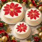 Three round white cakes decorated with large red poinsettia flowers and yellow centers, arranged on a marble surface with Christmas greenery, ribbons, and gold ornaments.