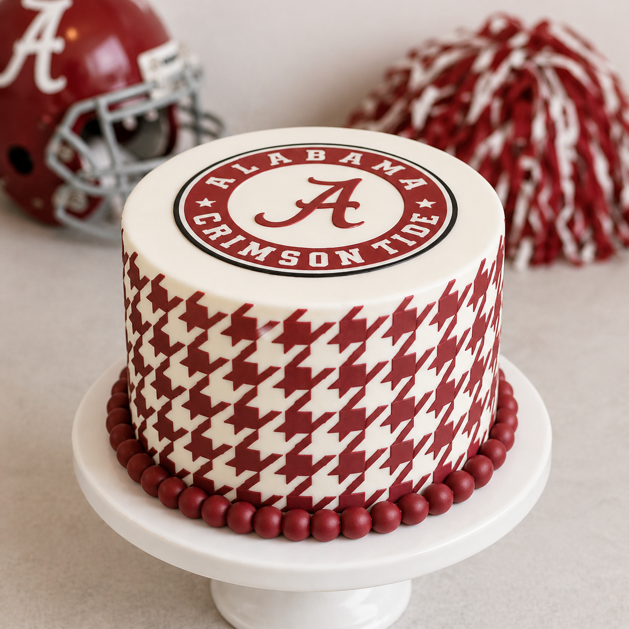 Cake with Alabama Crimson Tide design on a white stand, surrounded by football helmet and pom-pom.