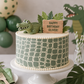Green birthday cake with alligator decoration and 'Happy Birthday Oliver!' sign, surrounded by green balloons and decor.