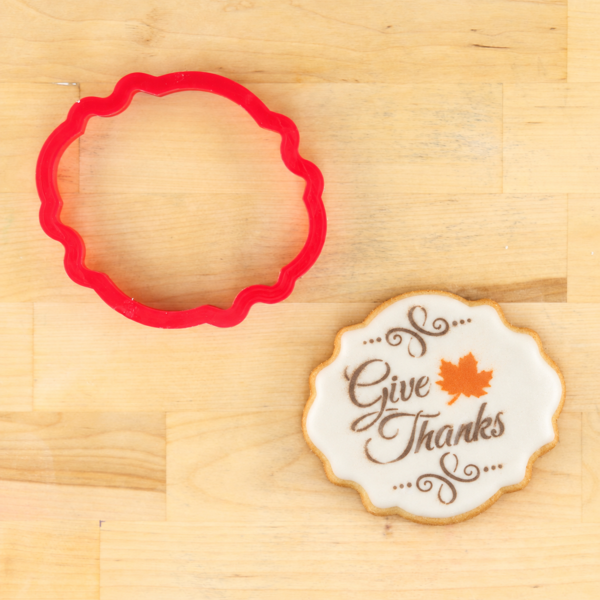 A red scalloped circular cookie cutter placed on a light wood surface next to a decorated sugar cookie with white icing, brown script text reading 'Give Thanks,' and an orange maple leaf.