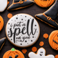 A styled flat-lay of Halloween-themed sugar cookies on a dark surface, featuring a round white cookie with black script reading 'i put a spell on you,' black witch hat cookies, orange pumpkin cookies, a ghost cookie, and orange confetti.