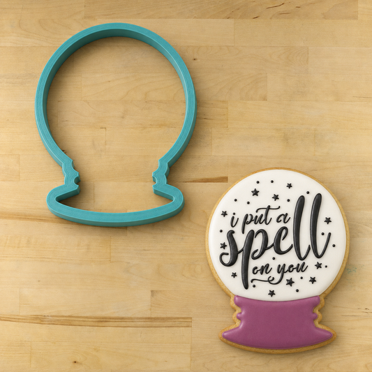 Snow globe cookie cutter used with the "I Put a Spell on You" Cookie Stencil applied to a white and purple iced cookie to make a crystal ball cookie for Hocus Pocus theme cookies