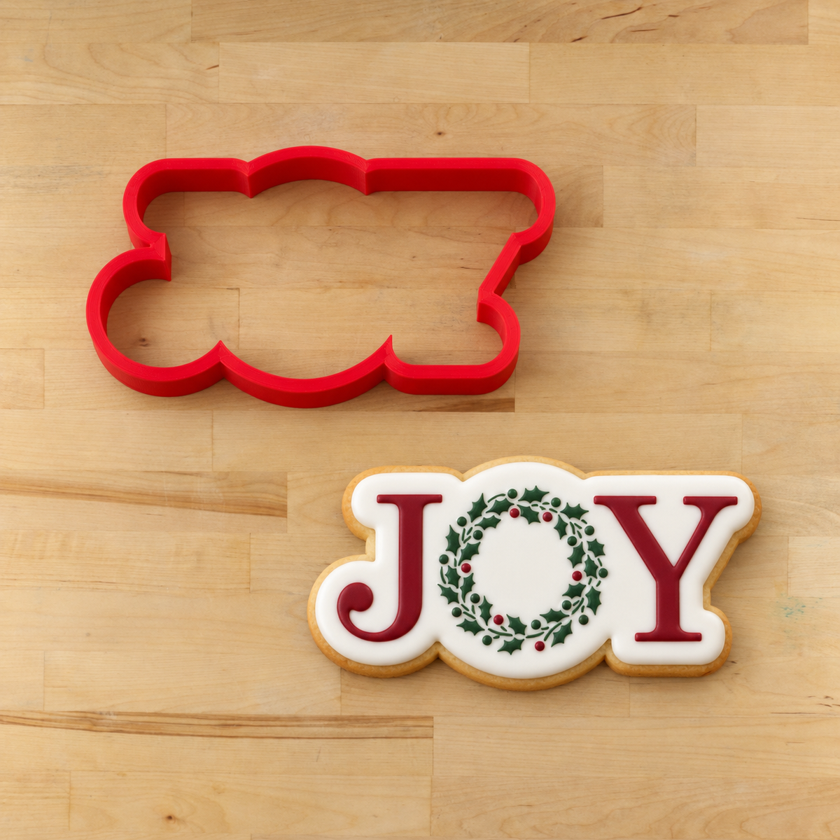 The JOY with Holiday Wreath Cookie Stencil  applied to a white flooded cookie lying next to its matching JOY cookie cutter.
