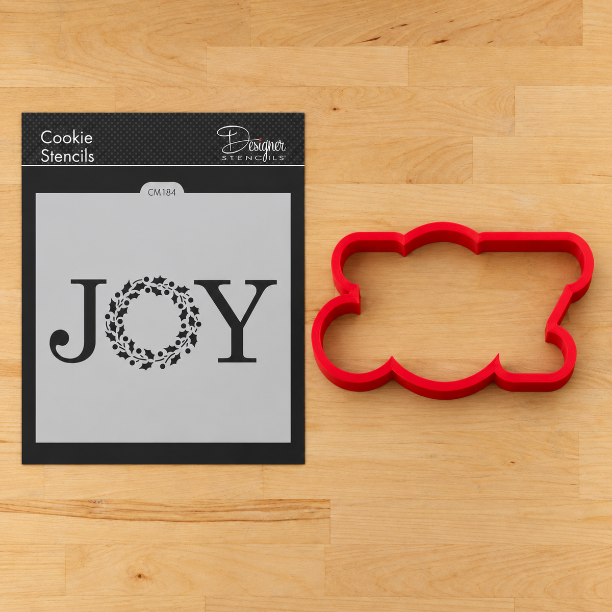 A red JOY-shaped cookie cutter and its JOY cookie stencil on a light wood background. The stencil shows the word JOY, with the letter O replaced by a Christmas wreath motif.
